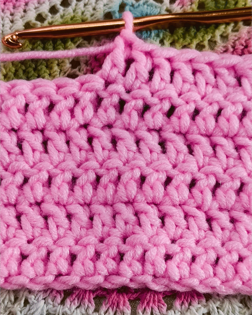A close-up of a pink swatch a tutorial to how to learn double crochet stitches, with a crochet hook inserted into the loops of the yarn.