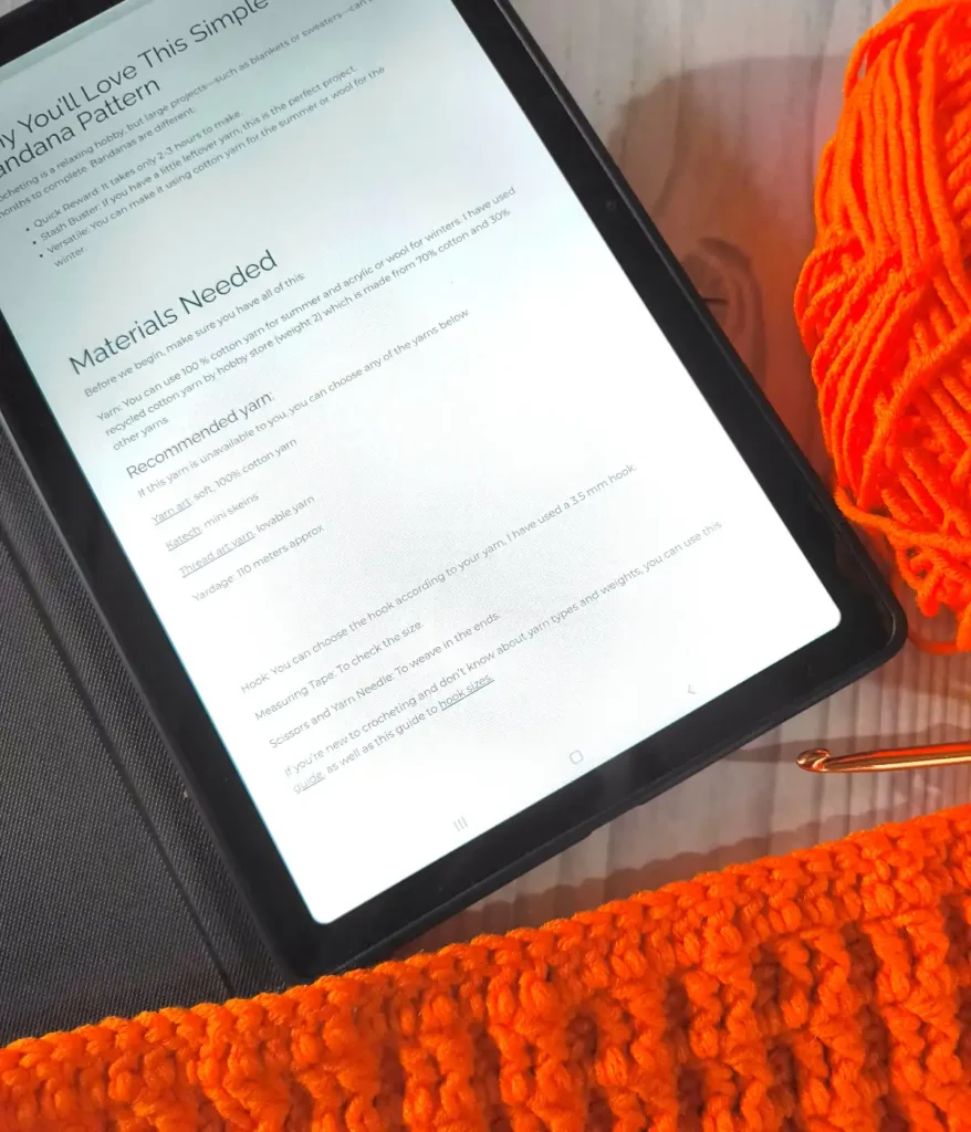 There is a tablet on a table showing a crochet pattern, along with an orange crochet swatch and orange yarn.