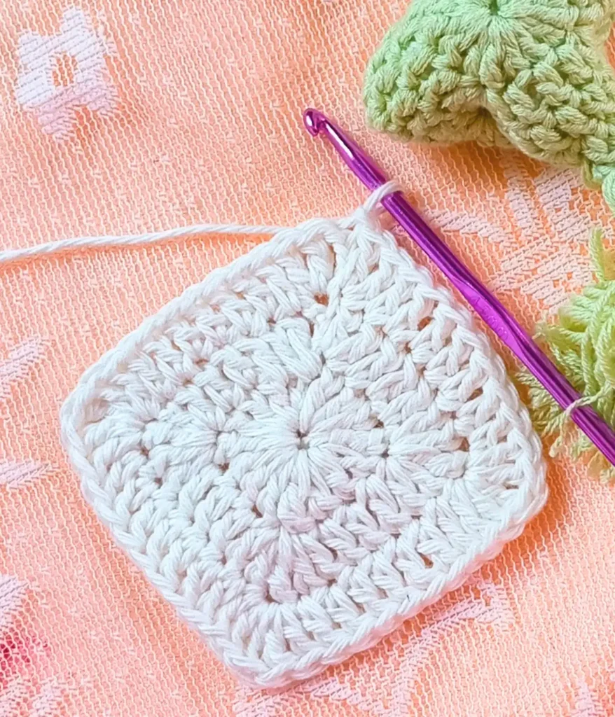 White color granny Square with a purple color crochet hook and peach color net background