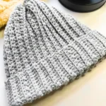 A grey coloured crocheted beanie hat is placed on a white background