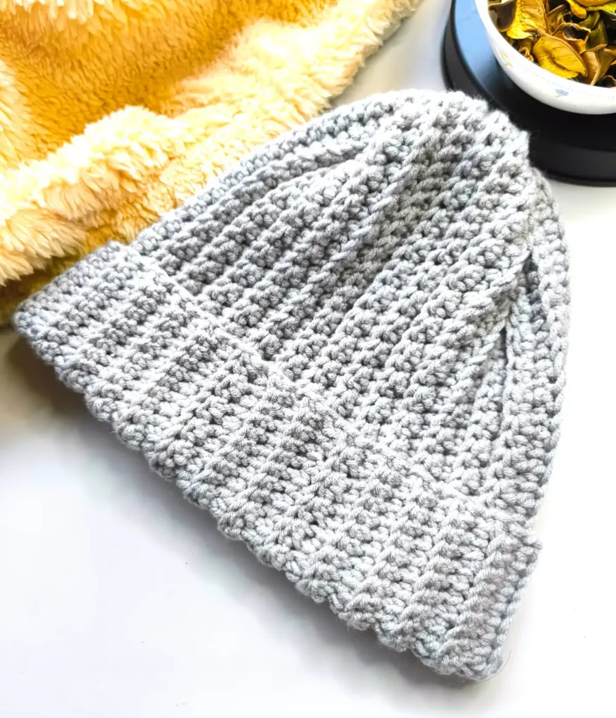 A grey coloured crocheted beanie hat is on the white table with fur and some dried leaves