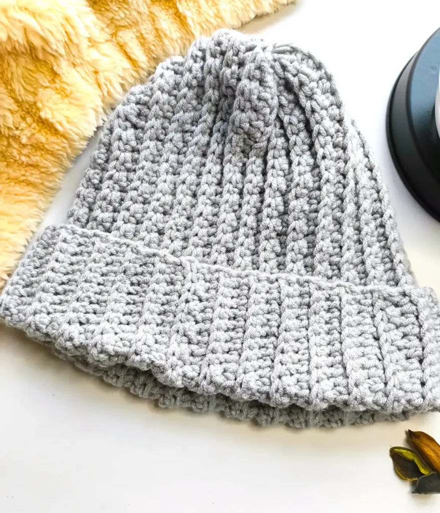A grey coloured crocheted beanie hat is placed on a white table with 3 dried leaves and a fur