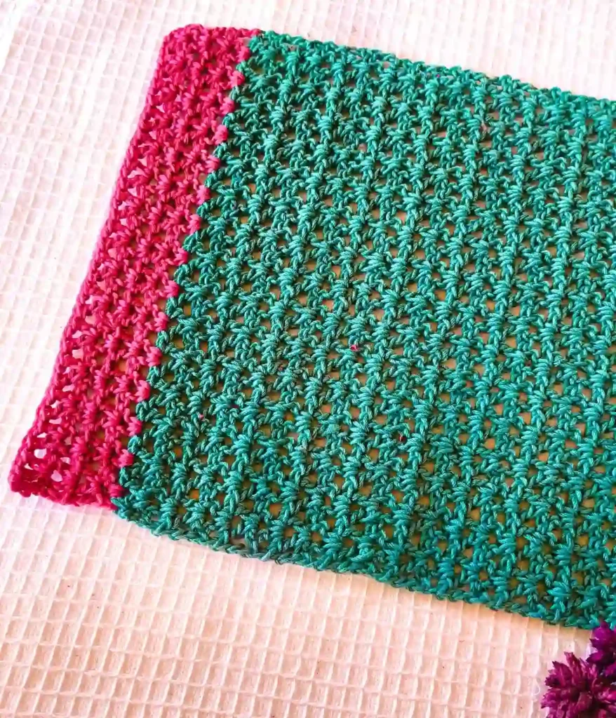 A green placemat made by crochet v stitch with bright pink small section, place at a white background along with some dried flowers